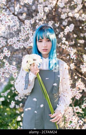 Moderne trendige Frau mit blauen Haaren hält Bouquet von frisch Blumen und Blick auf die Kamera, während im blühenden Frühling stehen Garten Stockfoto