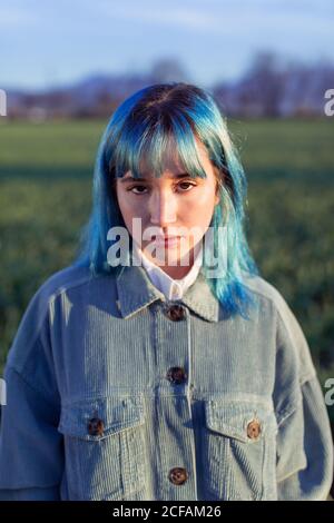 Nachdenklich junge Frau mit blauen Haaren Blick auf Kamera gekleidet In modischer Jacke im grünen Feld in sonnigen Abend stehen Stockfoto