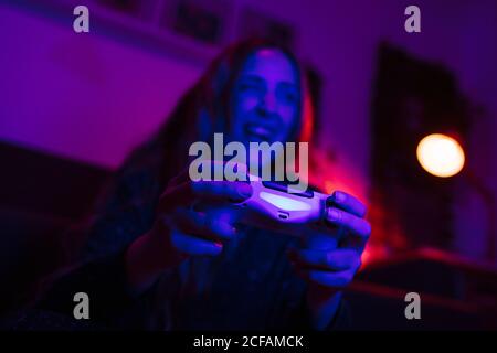 Nahaufnahme der Hände eines Smiley-Mädchens, das einen Controller hält und Videospiele spielt. Sie ist in einem Wohnzimmer mit bunten Lichtern in der Nacht. Stockfoto