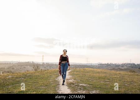 Ganzkörper der jungen Dame in stilvollen Casual Outfit zu Fuß Auf schmalem Weg zwischen Wiese mit grünem Gras und grau Himmel im Hintergrund Stockfoto