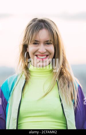 Moderne fröhliche blonde junge Frau in farbenfroher Jacke und grünem Neontop, die auf die Kamera mit abgelegenem Land auf dem Hintergrund schaut Stockfoto