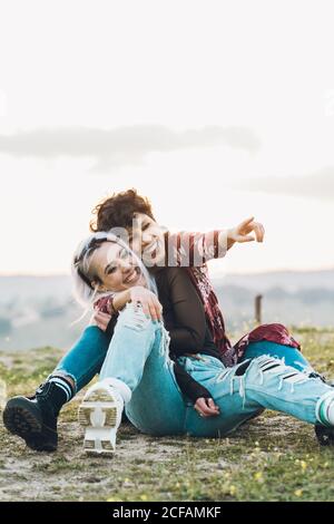 Lachend enge Freundinnen in stilvollen Casual Outfits sitzen auf grün Gras in Umarmung mit Himmel auf Hintergrund Stockfoto