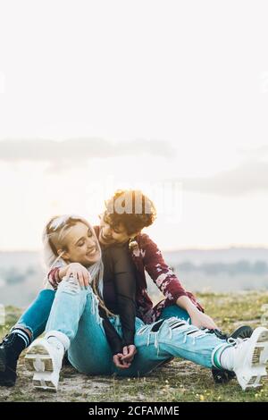 Lachend enge Freundinnen in stilvollen Casual Outfits sitzen auf grün Gras in Umarmung mit Himmel auf Hintergrund Stockfoto