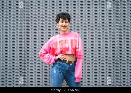 Selbstbewusste fröhliche ethnische schwarz haarige Dame in stilvollen rosa Sweatshirt Und Jeans stehen mit der Hand auf der Taille lächelnd weg in Vorderseite der grauen Wand Stockfoto