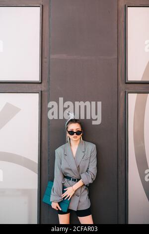 Elegante Geschäftsfrau mit modischer Sonnenbrille und Anzug, der Laptop hält und die Kamera an der Wand anschaut Stockfoto