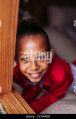 Von oben von niedlichen kleinen lockig behaarten ethnischen Mädchen mit Rote Schleife trägt rotes Kleid Blick auf Kamera und lächelnd Vor unscharfem Hintergrund Stockfoto