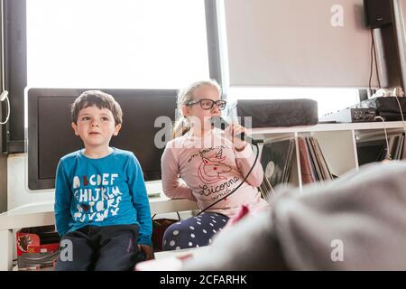 Mädchen in Gläsern singen Karaoke-Lied, während sie in der Nähe von Bruder in einem gemütlichen Zimmer am Wochenende zu Hause sitzen Stockfoto