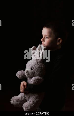 Studio-Aufnahme von ruhigen kleinen Jungen in lässigen Pullover umarmt Lieblings Plüsch grauen Hase beim Blick weg und posiert auf Schwarzer Hintergrund Stockfoto