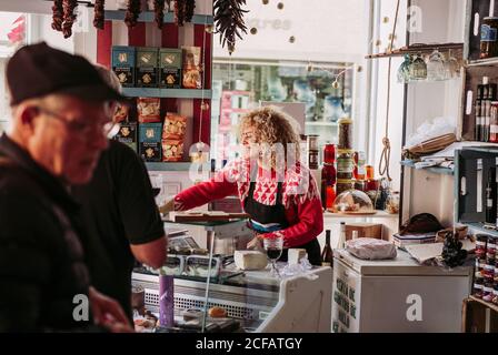 Senior Mann Auswahl Lebensmittel aus dem Gefrierschrank beim Kauf von Wein und Käse von Verkäuferin im gemütlichen kleinen Feinkostladen Stockfoto