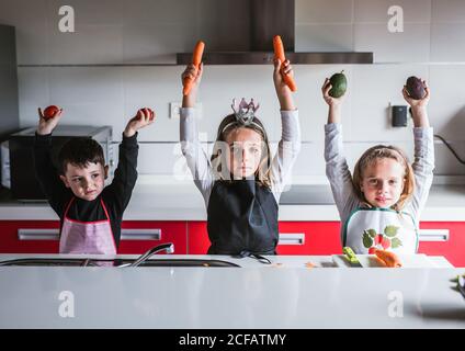 Kleine Mädchen und Jungen in Schürzen halten verschiedene frische Gemüse Mit erhobenen Händen und Blick auf die Kamera, während sie im Stehen Küche zu Hause zusammen Stockfoto