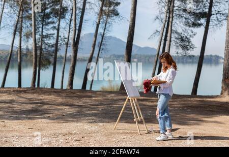 Seitenansicht der schönen jungen Dame im lässigen Outfit stehen In der Nähe Staffelei und Malerei während der Zeit in der Nähe von ruhigen See An sonnigen Tag auf dem Land Stockfoto
