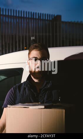 Erwachsener bärtiger Mann in Brille, der denkt und die Kamera anschaut Beim Stehen in der Nähe des Autos und halten Pappkartons am Abend Stockfoto