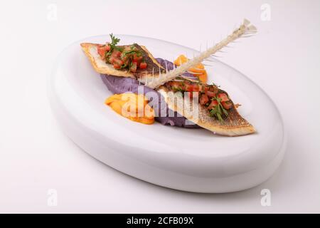 Wolfsbarschfilet mit violettem Kartoffelpüree, Karottenpüree und Tomatensalsa. Serviert auf weißem Hintergrund. Stockfoto