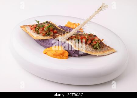 Wolfsbarschfilet mit violettem Kartoffelpüree, Karottenpüree und Tomatensalsa. Serviert auf weißem Hintergrund. Stockfoto