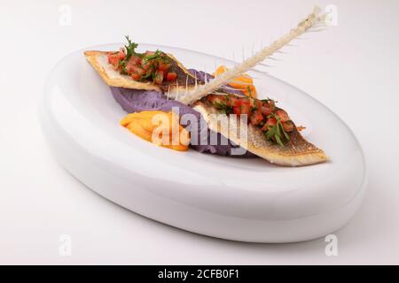 Wolfsbarschfilet mit violettem Kartoffelpüree, Karottenpüree und Tomatensalsa. Serviert auf weißem Hintergrund. Stockfoto