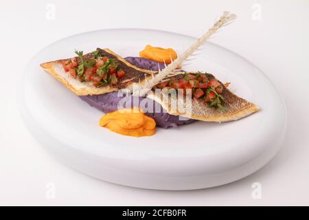 Wolfsbarschfilet mit violettem Kartoffelpüree, Karottenpüree und Tomatensalsa. Serviert auf weißem Hintergrund. Stockfoto