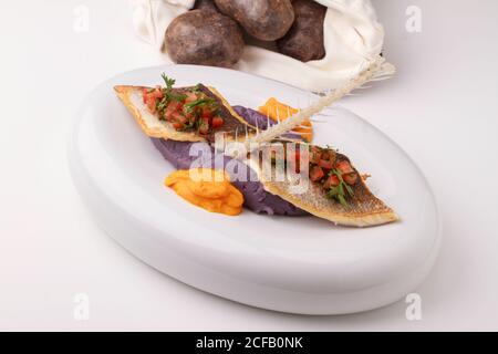 Wolfsbarschfilet mit violettem Kartoffelpüree, Karottenpüree und Tomatensalsa. Serviert auf weißem Hintergrund. Stockfoto