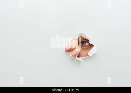 Blaues Auge von nicht erkennbaren Weibchen mit Fingern zum wegschauen Durch gerissene Loch in weißem Papier Karton Hintergrund Stockfoto