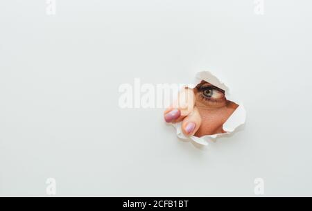 Blaues Auge von nicht erkennbaren Weibchen mit Fingern zum wegschauen Durch gerissene Loch in weißem Papier Karton Hintergrund Stockfoto