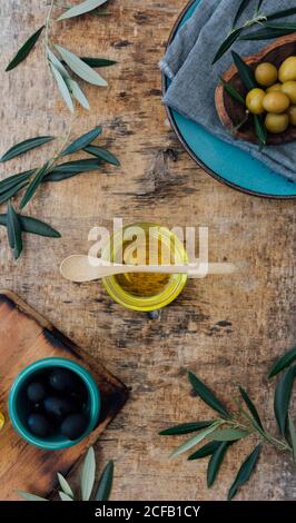 Von oben Glas mit Bio-Olivenöl mit Holzlöffel auf schäbigen grauen Tisch in der Nähe von frischem Grün platziert Oliven und Äste Stockfoto