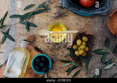 Von oben Glas mit Bio-Olivenöl mit Holzlöffel auf schäbigen grauen Tisch mit frischen Tomaten platziert Grüne und schwarze Oliven in der Nähe von Ästen Stockfoto