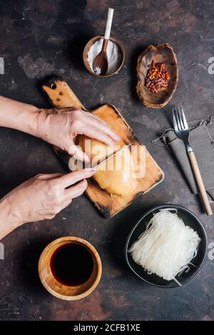 Von oben der Ernte anonyme Frau schneiden rohes Hühnerfilet auf Holz Schneidebrett während der Zubereitung von Nudeln für das Mittagessen Stockfoto