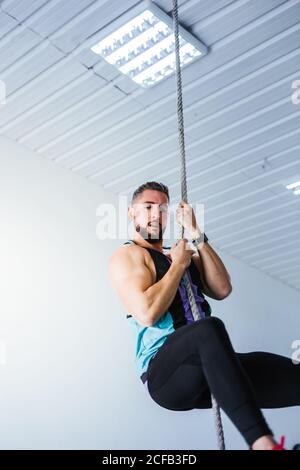 Von unten Seitenansicht des jungen athletischen Männchens in Sportbekleidung Durchführung von Seilklettern Übung während des Fitnesstrainings in modernen Fitnessraum Stockfoto