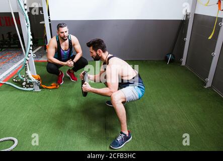High-Angle-Seitenansicht des fokussierten starken Sportlers beim Hocken Übung mit Gewichtscheibe beim Training im Fitnessstudio mit Trainer Stockfoto