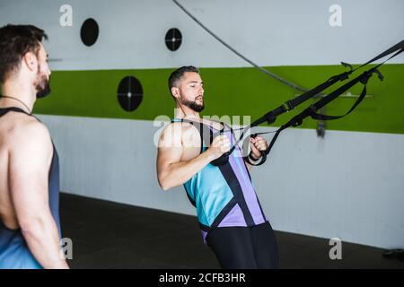 Seitenansicht des jungen muskulösen Mannes im Sportswear Training mit Elastische Widerstandsbänder beim intensiven Training mit Freund in modern Fitnessraum Stockfoto