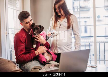Fröhliche Schwangere und Ehemann verbringen Zeit zu Hause und Spaß beim Spielen mit Hund in rosa Kostüm gekleidet Stockfoto