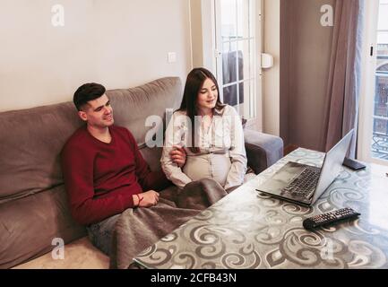 Junger Mann im roten Pullover sitzt mit schwangeren Frau in Beige Bluse auf Sofa und berühren Baby Bump während der Ausgaben Gemeinsam am Computer Wintertag zu Hause Stockfoto
