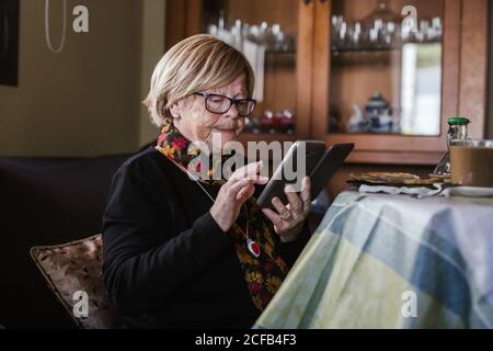 Ältere Frau schaut und berührt den Bildschirm des Smartphones, während sie auf dem Sofa im Wohnzimmer sitzt Stockfoto
