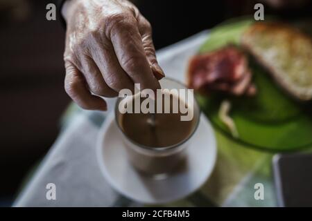 Hand einer älteren Frau, die den Löffel in der Tasse hält, während sie beim Frühstück Kaffee mischt Stockfoto