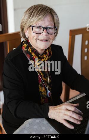 Ältere Frau in einer Brille, die die Kamera beim Surfen im Laptop anschaut, während sie am Tisch in einem gemütlichen Zuhause sitzt Stockfoto