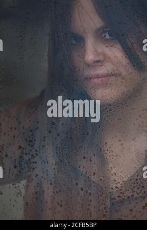 Porträt einer schönen Frau, die am regnerischen Tag aus dem nassen Fenster schaut und die Kamera anschaut Stockfoto