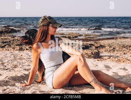 Positive junge Frau in legerer Kleidung und Mütze sitzt am Sandstrand mit dem Hund Rottweiler, während sie im Sommer an einem sonnigen Tag Zeit verbringt Stockfoto