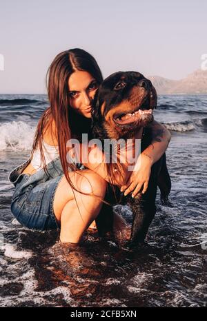 Glückliche junge Frau in legerer Kleidung kauerte sich mit einem großen schwarzen Hund im Wasser an der Küste und schaute auf die Kamera, während sie am Abend Zeit verbrachte Stockfoto