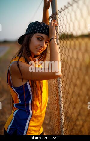 Seitenansicht der jungen sportlichen Dame in stilvoller Sportswear und Kappe steht in der Nähe von Metallzaun und Blick auf die Kamera während Zeit auf der Straße verbringen Stockfoto