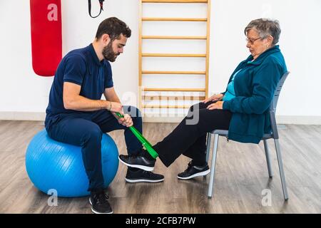 Seitenansicht eines männlichen persönlichen Trainers, der auf einem blauen Übungsball sitzt, während er ein grünes Gummiband am Knöchel einer alten Frau im Fitnessstudio verwendet Stockfoto