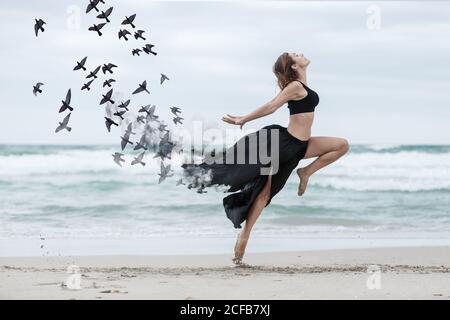 Seitenansicht der barfuß schlanken Frau, die auf nassem Sand tanzt In der Nähe winkende Meer, während schwarze Vögel fliegen vom Rock Stockfoto