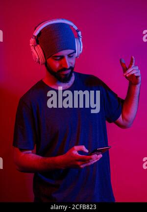 Junger bärtiger Mann in Kopfhörern auf dem Smartphone und gestikulierenden shaka melden Sie sich an, während Sie unter rotem Licht Musik hören Stockfoto