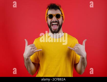 Bärtig trendy Mann in gelbem Outfit zeigt Zunge und Gestik shaka-Schild vor hellrotem Hintergrund Stockfoto