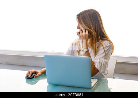 Frau, die am Glastisch am Fenster sitzt und auf dem Smartphone spricht Durchsuchen von Laptops und Verwenden einer drahtlosen Maus im Büro Stockfoto