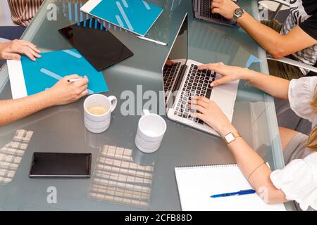 Von oben Ernte Mitarbeiter sitzen am Glastisch mit Kaffee Tassen tippen auf Laptops mit Gadgets und Notizen Stockfoto