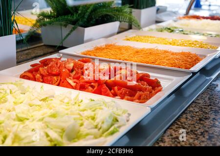 Frisch gehackte Kohl Tomaten Karotten Mais und anderes Gemüse in Vitrinen auf verschwommenem Hintergrund von Topfpflanzen in heller Kantine Stockfoto