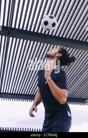 Von unten Seitenansicht des jungen bärtigen Fußballs im Dunkeln Blaue Sportswear, die beim Training auf dem überdachten Spielplatz den Ball steuert Stockfoto