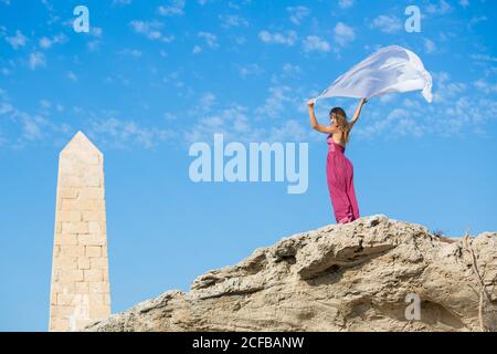 Von unten schlanke junge Dame im Kleid mit Schärpe in Hochgepuppte Hände posieren auf Stein in der Nähe von Felsdenkmal und blau Himmel Stockfoto