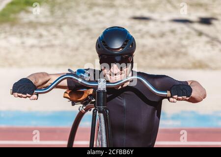 Portrait von glücklichen Mann in schwarz activewear und Helm suchen Bei der Kamera, während Sie mit Retro Penny Farthing Fahrrad auf stehen Rennbahn am Sportplatz Stockfoto