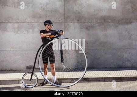 Ruhiger entschlossener Mann in schwarzer Aktivbekleidung und Helm, der wegschaut Auf Asphaltstraße stehend und auf hohem Rad gelehnt Fahrrad gegen Betonwand an sonnigen Tagen Stockfoto