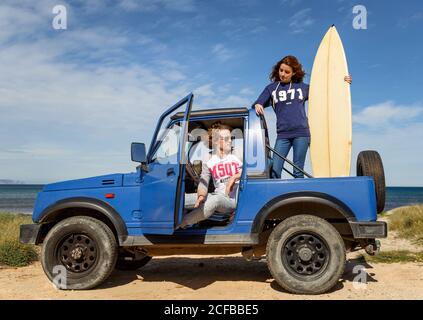 Junger energischer Mann mit Surfbrett und attraktiver Frau, die an einem hellen Tag auf dem Rücken eines blauen Offroad-Sportboards steht Stockfoto